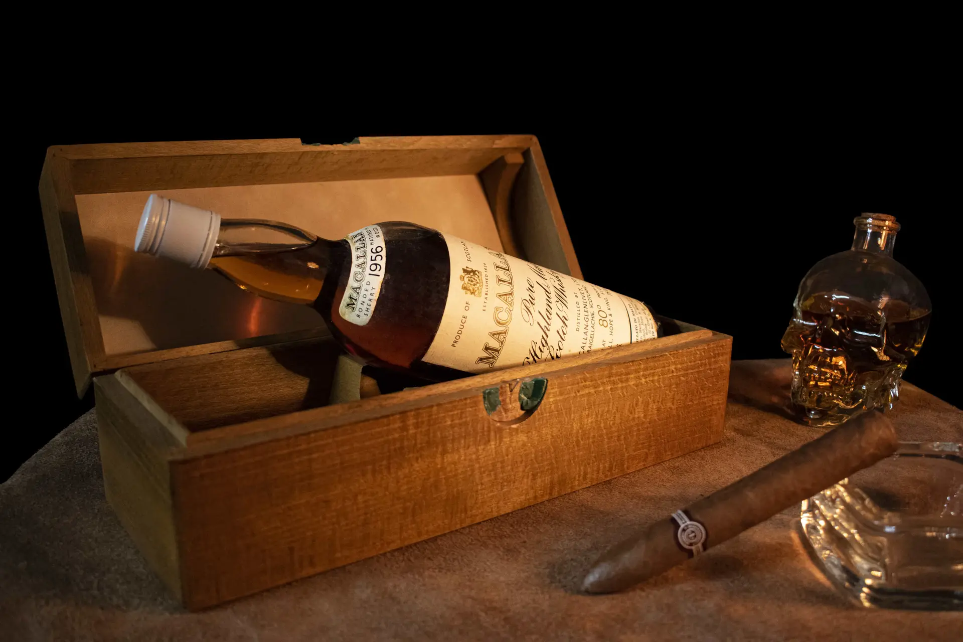 Bottiglia di whisky ambrato in primo piano, con un bicchiere di whisky e un sigaro su un tavolo di legno rustico. Un libro antico aperto sullo sfondo aggiunge un tocco di eleganza.